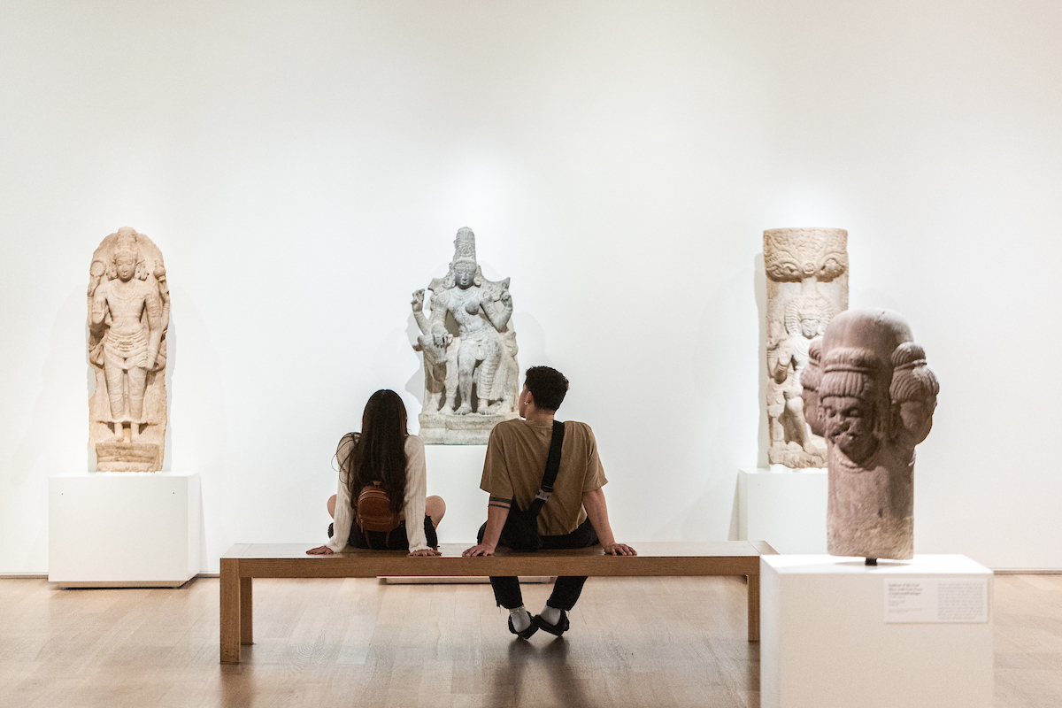 Two students sit on a bench and look at ancient South Asian sculptures in the Art Institute of Chicago.