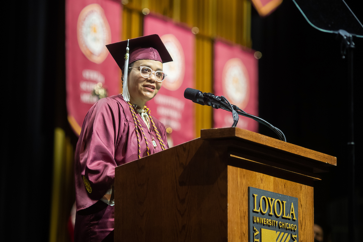 Woman wearing glasses and academic regalia speaks at a podium.